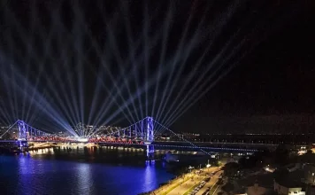 Vista aérea mostra o público reunido na Beira-Mar Continental durante as comemorações do centenário da Ponte Hercílio Luz, em Florianópolis, com palco de shows, iluminação especial e feixes de luz sobre a ponte durante evento noturno.