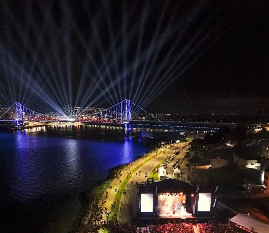 Vista aérea mostra o público reunido na Beira-Mar Continental durante as comemorações do centenário da Ponte Hercílio Luz, em Florianópolis, com palco de shows, iluminação especial e feixes de luz sobre a ponte durante evento noturno.