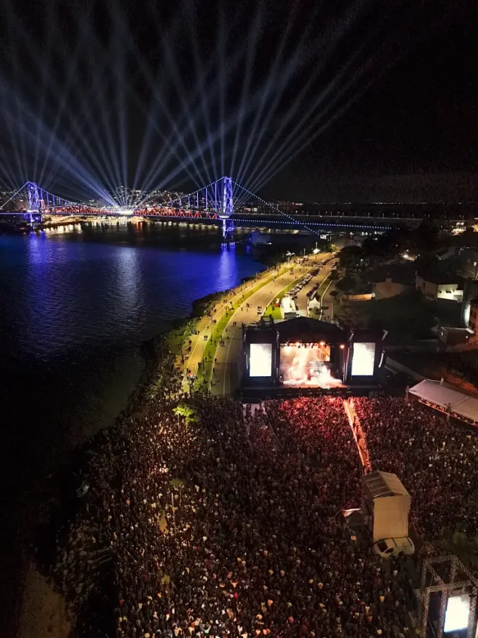 Vista aérea mostra o público reunido na Beira-Mar Continental durante as comemorações do centenário da Ponte Hercílio Luz, em Florianópolis, com palco de shows, iluminação especial e feixes de luz sobre a ponte durante evento noturno.