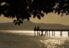 Silhueta de duas pessoas em um píer sobre o mar ao entardecer, com reflexo do sol na água e montanhas ao fundo, ilustrando cenário de tempo firme e temperaturas amenas antes da chegada de frente fria.