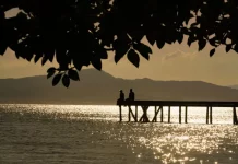 Silhueta de duas pessoas em um píer sobre o mar ao entardecer, com reflexo do sol na água e montanhas ao fundo, ilustrando cenário de tempo firme e temperaturas amenas antes da chegada de frente fria.