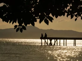 Silhueta de duas pessoas em um píer sobre o mar ao entardecer, com reflexo do sol na água e montanhas ao fundo, ilustrando cenário de tempo firme e temperaturas amenas antes da chegada de frente fria.