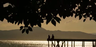 Silhueta de duas pessoas em um píer sobre o mar ao entardecer, com reflexo do sol na água e montanhas ao fundo, ilustrando cenário de tempo firme e temperaturas amenas antes da chegada de frente fria.
