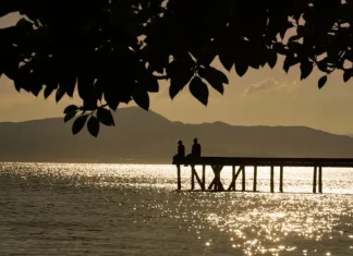 Silhueta de duas pessoas em um píer sobre o mar ao entardecer, com reflexo do sol na água e montanhas ao fundo, ilustrando cenário de tempo firme e temperaturas amenas antes da chegada de frente fria.