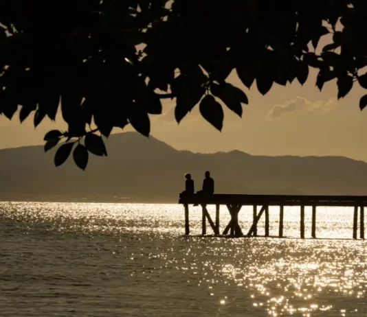Silhueta de duas pessoas em um píer sobre o mar ao entardecer, com reflexo do sol na água e montanhas ao fundo, ilustrando cenário de tempo firme e temperaturas amenas antes da chegada de frente fria.