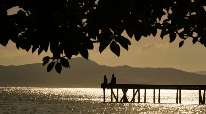 Frente fria provoca temporais e queda de temperatura no fim de semana Silhueta de duas pessoas em um píer sobre o mar ao entardecer, com reflexo do sol na água e montanhas ao fundo, ilustrando cenário de tempo firme e temperaturas amenas antes da chegada de frente fria.