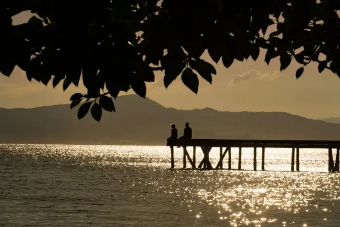 Silhueta de duas pessoas em um píer sobre o mar ao entardecer, com reflexo do sol na água e montanhas ao fundo, ilustrando cenário de tempo firme e temperaturas amenas antes da chegada de frente fria.