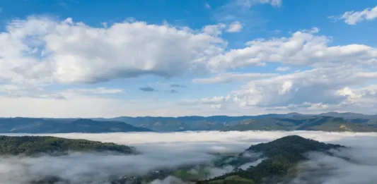 Frio intenso marca início da semana em Santa Catarina Vista aérea de uma região montanhosa de Santa Catarina coberta por nevoeiro e nuvens baixas ao amanhecer. A imagem mostra vegetação verde, morros e o céu parcialmente nublado, ilustrando o frio intenso e as condições típicas da massa de ar polar que atua no estado.