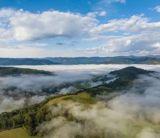 Frio intenso marca início da semana em Santa Catarina Vista aérea de uma região montanhosa de Santa Catarina coberta por nevoeiro e nuvens baixas ao amanhecer. A imagem mostra vegetação verde, morros e o céu parcialmente nublado, ilustrando o frio intenso e as condições típicas da massa de ar polar que atua no estado.