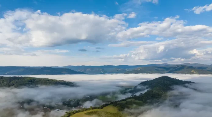 Frio intenso marca início da semana em Santa Catarina Vista aérea de uma região montanhosa de Santa Catarina coberta por nevoeiro e nuvens baixas ao amanhecer. A imagem mostra vegetação verde, morros e o céu parcialmente nublado, ilustrando o frio intenso e as condições típicas da massa de ar polar que atua no estado.
