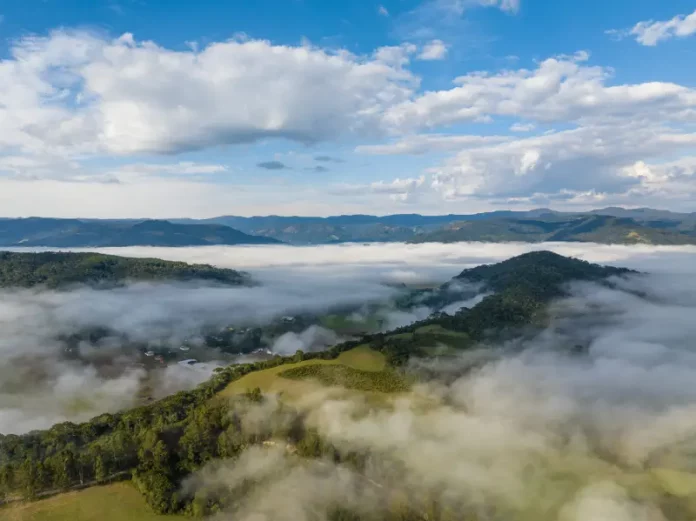 frio-intenso Vista aérea de uma região montanhosa de Santa Catarina coberta por nevoeiro e nuvens baixas ao amanhecer. A imagem mostra vegetação verde, morros e o céu parcialmente nublado, ilustrando o frio intenso e as condições típicas da massa de ar polar que atua no estado.