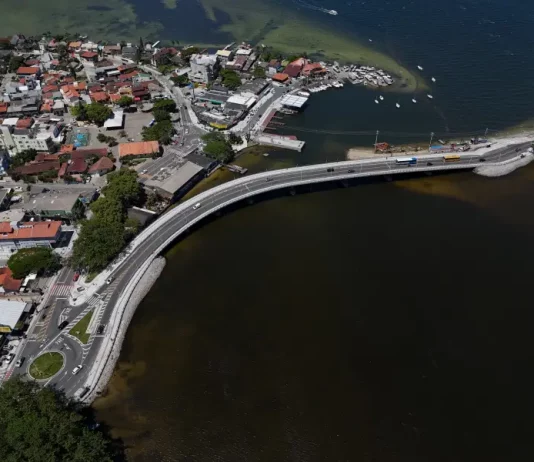 Imagem aérea da nova ponte da Lagoa da Conceição, em Florianópolis, mostrando a estrutura sobre a água, o tráfego de veículos e a ligação viária entre as margens, destacando sua função de melhorar a mobilidade e o acesso às praias do Leste.