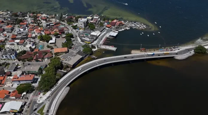 Imagem aérea da nova ponte da Lagoa da Conceição, em Florianópolis, mostrando a estrutura sobre a água, o tráfego de veículos e a ligação viária entre as margens, destacando sua função de melhorar a mobilidade e o acesso às praias do Leste.