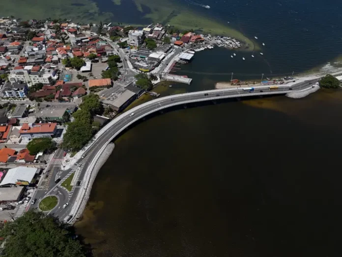 Imagem aérea da nova ponte da Lagoa da Conceição, em Florianópolis, mostrando a estrutura sobre a água, o tráfego de veículos e a ligação viária entre as margens, destacando sua função de melhorar a mobilidade e o acesso às praias do Leste.