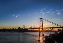 Ponte suspensa iluminada pelo pôr do sol em Florianópolis, com a Ponte Hercílio Luz atravessando o mar e conectando a ilha ao continente; ao fundo, silhueta da cidade sob céu com tons alaranjados e azuis.