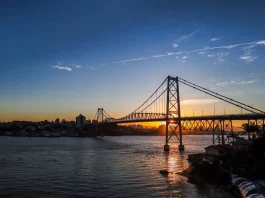 Ponte suspensa iluminada pelo pôr do sol em Florianópolis, com a Ponte Hercílio Luz atravessando o mar e conectando a ilha ao continente; ao fundo, silhueta da cidade sob céu com tons alaranjados e azuis.