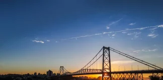 Ponte suspensa iluminada pelo pôr do sol em Florianópolis, com a Ponte Hercílio Luz atravessando o mar e conectando a ilha ao continente; ao fundo, silhueta da cidade sob céu com tons alaranjados e azuis.