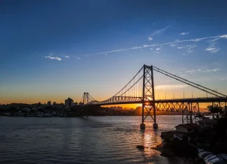 Ponte suspensa iluminada pelo pôr do sol em Florianópolis, com a Ponte Hercílio Luz atravessando o mar e conectando a ilha ao continente; ao fundo, silhueta da cidade sob céu com tons alaranjados e azuis.
