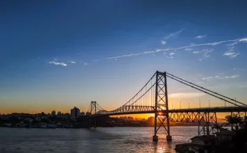 Ponte suspensa iluminada pelo pôr do sol em Florianópolis, com a Ponte Hercílio Luz atravessando o mar e conectando a ilha ao continente; ao fundo, silhueta da cidade sob céu com tons alaranjados e azuis.