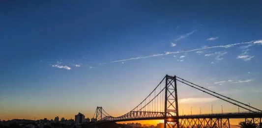 Ponte suspensa iluminada pelo pôr do sol em Florianópolis, com a Ponte Hercílio Luz atravessando o mar e conectando a ilha ao continente; ao fundo, silhueta da cidade sob céu com tons alaranjados e azuis.
