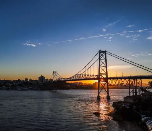 Ponte suspensa iluminada pelo pôr do sol em Florianópolis, com a Ponte Hercílio Luz atravessando o mar e conectando a ilha ao continente; ao fundo, silhueta da cidade sob céu com tons alaranjados e azuis.