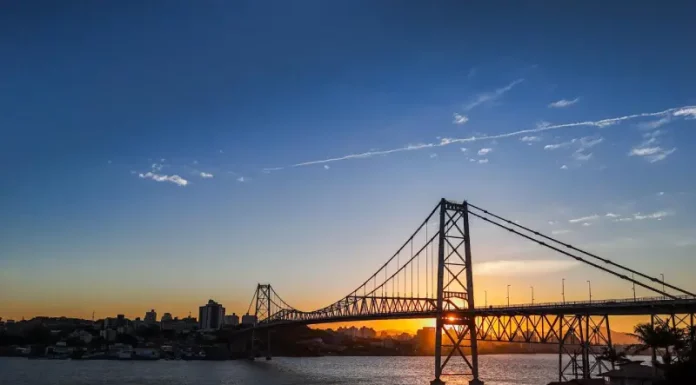 Ponte suspensa iluminada pelo pôr do sol em Florianópolis, com a Ponte Hercílio Luz atravessando o mar e conectando a ilha ao continente; ao fundo, silhueta da cidade sob céu com tons alaranjados e azuis.