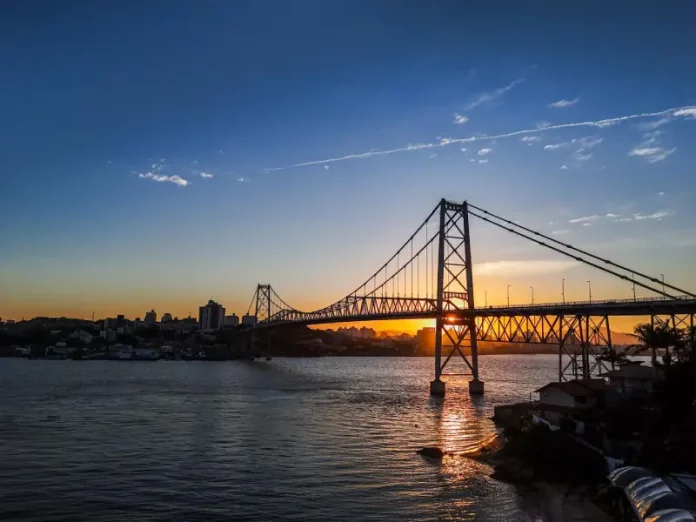 Ponte suspensa iluminada pelo pôr do sol em Florianópolis, com a Ponte Hercílio Luz atravessando o mar e conectando a ilha ao continente; ao fundo, silhueta da cidade sob céu com tons alaranjados e azuis.