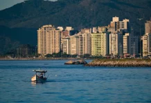 Vista de uma cidade litorânea em Santa Catarina com prédios altos à beira-mar, montanhas ao fundo e um pequeno barco sobre águas calmas, sob céu ensolarado — imagem que ilustra a previsão de calor e tempo firme no início do feriado do Dia do Trabalho antes da chegada de instabilidade e frente fria no fim de semana.