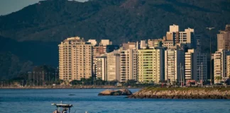 Vista de uma cidade litorânea em Santa Catarina com prédios altos à beira-mar, montanhas ao fundo e um pequeno barco sobre águas calmas, sob céu ensolarado — imagem que ilustra a previsão de calor e tempo firme no início do feriado do Dia do Trabalho antes da chegada de instabilidade e frente fria no fim de semana.