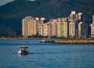 Vista de uma cidade litorânea em Santa Catarina com prédios altos à beira-mar, montanhas ao fundo e um pequeno barco sobre águas calmas, sob céu ensolarado — imagem que ilustra a previsão de calor e tempo firme no início do feriado do Dia do Trabalho antes da chegada de instabilidade e frente fria no fim de semana.