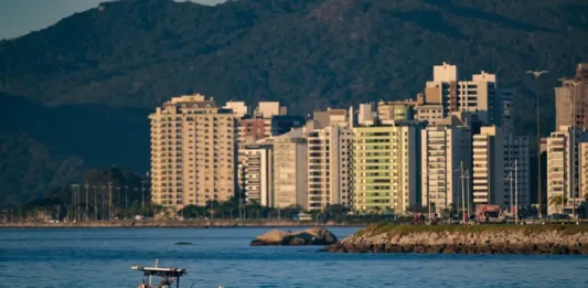 Vista de uma cidade litorânea em Santa Catarina com prédios altos à beira-mar, montanhas ao fundo e um pequeno barco sobre águas calmas, sob céu ensolarado — imagem que ilustra a previsão de calor e tempo firme no início do feriado do Dia do Trabalho antes da chegada de instabilidade e frente fria no fim de semana.