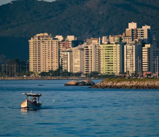 Vista de uma cidade litorânea em Santa Catarina com prédios altos à beira-mar, montanhas ao fundo e um pequeno barco sobre águas calmas, sob céu ensolarado — imagem que ilustra a previsão de calor e tempo firme no início do feriado do Dia do Trabalho antes da chegada de instabilidade e frente fria no fim de semana.