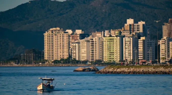 Dia do Trabalho será de calor, mas frente fria muda o tempo no fim de semana Vista de uma cidade litorânea em Santa Catarina com prédios altos à beira-mar, montanhas ao fundo e um pequeno barco sobre águas calmas, sob céu ensolarado — imagem que ilustra a previsão de calor e tempo firme no início do feriado do Dia do Trabalho antes da chegada de instabilidade e frente fria no fim de semana.