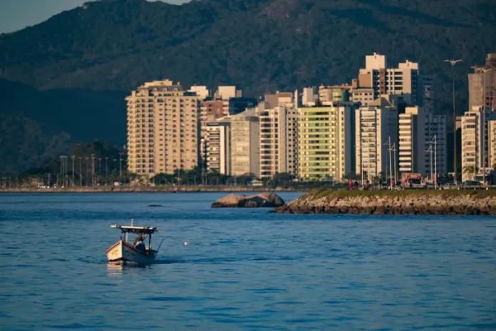 Vista de uma cidade litorânea em Santa Catarina com prédios altos à beira-mar, montanhas ao fundo e um pequeno barco sobre águas calmas, sob céu ensolarado — imagem que ilustra a previsão de calor e tempo firme no início do feriado do Dia do Trabalho antes da chegada de instabilidade e frente fria no fim de semana.