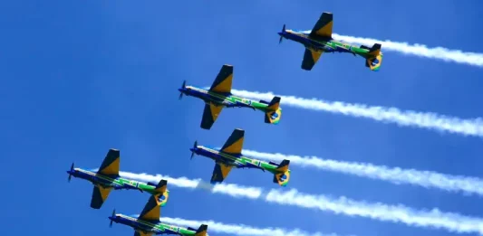 Seis aeronaves da Esquadrilha da Fumaça realizam voo em formação no céu azul, deixando rastros de fumaça branca. A imagem ilustra a apresentação aérea anunciada para Florianópolis e destaca o caráter de espetáculo visual e demonstração de precisão da Força Aérea Brasileira para o público.