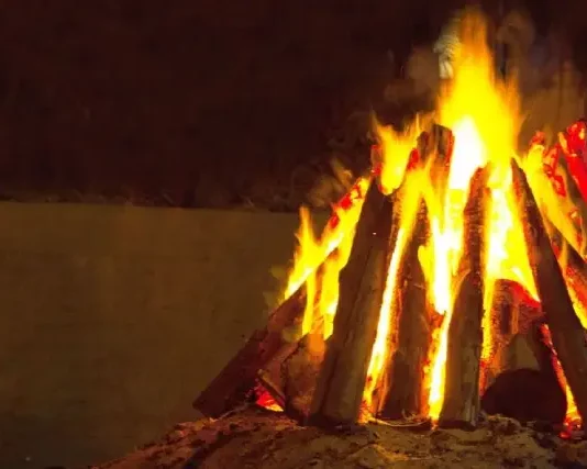 Fogueira acesa com toras de madeira empilhadas em formato triangular durante a noite. A imagem ilustra o tema das ocorrências com fogueiras e os cuidados necessários para prevenir queimaduras e incêndios durante as festas juninas.