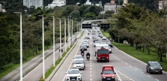 Florianópolis aprova 34 pontos de recarga para veículos elétricos em espaços públicos. Vista de uma avenida movimentada em Florianópolis, com carros circulando em múltiplas faixas e área urbana ao fundo. A imagem ilustra a expansão da mobilidade urbana e a implantação de pontos de recarga para veículos elétricos em espaços públicos da cidade.