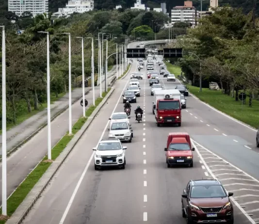 Florianópolis aprova 34 pontos de recarga para veículos elétricos em espaços públicos. Vista de uma avenida movimentada em Florianópolis, com carros circulando em múltiplas faixas e área urbana ao fundo. A imagem ilustra a expansão da mobilidade urbana e a implantação de pontos de recarga para veículos elétricos em espaços públicos da cidade.