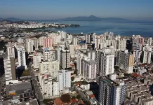 Vista aérea da região central e da área litorânea de São José, com prédios residenciais e comerciais próximos à Baía Sul. A imagem ilustra o crescimento urbano do município e a regulamentação da outorga onerosa, mecanismo que permite ampliar o potencial construtivo e alterar o uso de imóveis mediante contrapartida financeira.