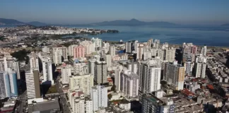 Vista aérea da região central e da área litorânea de São José, com prédios residenciais e comerciais próximos à Baía Sul. A imagem ilustra o crescimento urbano do município e a regulamentação da outorga onerosa, mecanismo que permite ampliar o potencial construtivo e alterar o uso de imóveis mediante contrapartida financeira.
