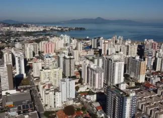 Vista aérea da região central e da área litorânea de São José, com prédios residenciais e comerciais próximos à Baía Sul. A imagem ilustra o crescimento urbano do município e a regulamentação da outorga onerosa, mecanismo que permite ampliar o potencial construtivo e alterar o uso de imóveis mediante contrapartida financeira.