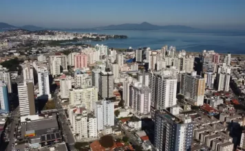 Vista aérea da região central e da área litorânea de São José, com prédios residenciais e comerciais próximos à Baía Sul. A imagem ilustra o crescimento urbano do município e a regulamentação da outorga onerosa, mecanismo que permite ampliar o potencial construtivo e alterar o uso de imóveis mediante contrapartida financeira.