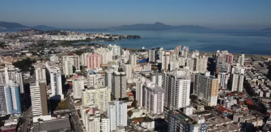 Vista aérea da região central e da área litorânea de São José, com prédios residenciais e comerciais próximos à Baía Sul. A imagem ilustra o crescimento urbano do município e a regulamentação da outorga onerosa, mecanismo que permite ampliar o potencial construtivo e alterar o uso de imóveis mediante contrapartida financeira.