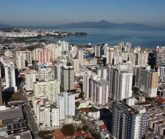Vista aérea da região central e da área litorânea de São José, com prédios residenciais e comerciais próximos à Baía Sul. A imagem ilustra o crescimento urbano do município e a regulamentação da outorga onerosa, mecanismo que permite ampliar o potencial construtivo e alterar o uso de imóveis mediante contrapartida financeira.