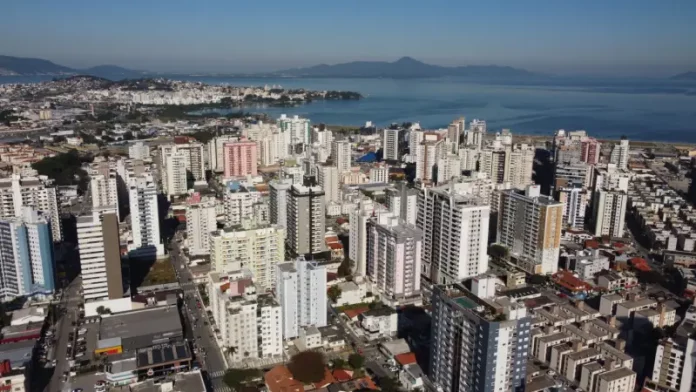 Vista aérea da região central e da área litorânea de São José, com prédios residenciais e comerciais próximos à Baía Sul. A imagem ilustra o crescimento urbano do município e a regulamentação da outorga onerosa, mecanismo que permite ampliar o potencial construtivo e alterar o uso de imóveis mediante contrapartida financeira.