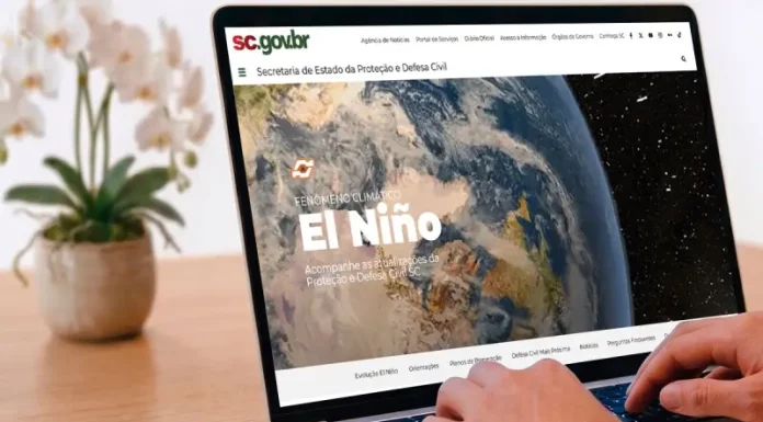 Pessoa utiliza um notebook exibindo a página especial “Fenômeno Climático El Niño” no portal da Secretaria de Estado da Proteção e Defesa Civil de Santa Catarina (SDC/SC). A imagem ilustra o acesso à plataforma criada para divulgar informações atualizadas, orientações de prevenção, documentos oficiais e conteúdos educativos sobre os impactos do El Niño e as ações de preparação da Defesa Civil no estado.