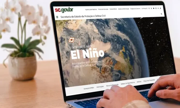 Pessoa utiliza um notebook exibindo a página especial “Fenômeno Climático El Niño” no portal da Secretaria de Estado da Proteção e Defesa Civil de Santa Catarina (SDC/SC). A imagem ilustra o acesso à plataforma criada para divulgar informações atualizadas, orientações de prevenção, documentos oficiais e conteúdos educativos sobre os impactos do El Niño e as ações de preparação da Defesa Civil no estado.