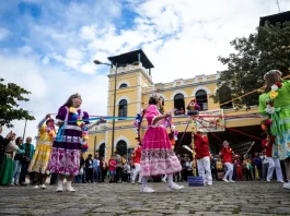 Participantes vestidos com trajes coloridos realizam uma apresentação cultural em frente ao Mercado Público de Florianópolis durante uma celebração do Dia do Manezinho. A imagem ilustra a valorização das tradições, da cultura popular e das manifestações folclóricas que fazem parte da identidade da cidade.