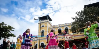 Participantes vestidos com trajes coloridos realizam uma apresentação cultural em frente ao Mercado Público de Florianópolis durante uma celebração do Dia do Manezinho. A imagem ilustra a valorização das tradições, da cultura popular e das manifestações folclóricas que fazem parte da identidade da cidade.