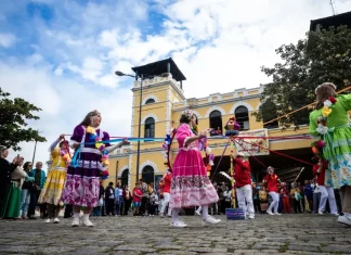 Participantes vestidos com trajes coloridos realizam uma apresentação cultural em frente ao Mercado Público de Florianópolis durante uma celebração do Dia do Manezinho. A imagem ilustra a valorização das tradições, da cultura popular e das manifestações folclóricas que fazem parte da identidade da cidade.